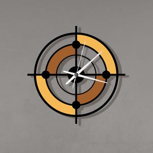 Puede incluir: Un reloj de pared moderno con un diseño de diana. El reloj presenta un marco negro con círculos concéntricos en tonos marrones y amarillos. Las manecillas del reloj son blancas y el reloj está montado en una pared gris.