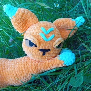 May include: A crocheted orange and turquoise plush toy cat with a V-shaped design on its forehead. The cat is lying on a bed of green grass.