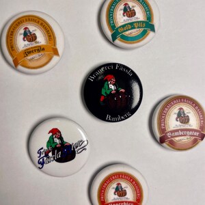 Könnte beinhalten: Fünf runde, bunte Knöpfe mit Bildern eines Zwergs, der ein Bierfass hält. Die Knöpfe zeigen den Text "Privatbrauerei Fässla Bamberg", "Zwergla", "Gold-Pils", "Brauerei Fässla Bamberg", "Bambergator", "Fassla Bier" und "Lagerbier".