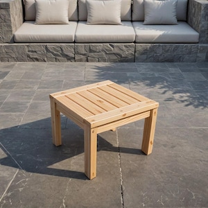 May include: A small, square wooden side table with a slatted top and four sturdy legs. The table is made of light-coloured wood and is positioned on a stone patio. The table is approximately 46 cm square and 41 cm high.