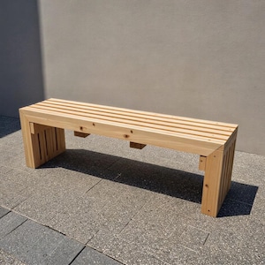 May include: A light-colored wooden bench with a slatted seat and rectangular legs. The bench is made of natural wood and is positioned outdoors on a stone surface. The bench is approximately 60 inches long.