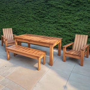 Puede incluir: Un juego de comedor de exterior de madera con una mesa rectangular y dos sillas. La mesa y las sillas están hechas de madera natural con un acabado marrón claro. El conjunto está colocado en un patio de color gris claro.