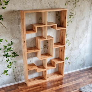 May include: A tall, wooden bookshelf with an open, geometric design. The natural wood tones of the shelves contrast with the light-colored wall and dark wood floor. The bookshelf has multiple cubbies and shelves of varying sizes.