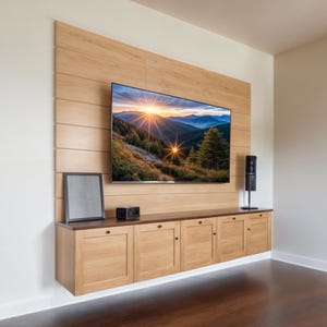 Puede incluir: Una unidad de pared de madera con un televisor de pantalla plana que muestra una imagen de paisaje. La unidad tiene un estante con un altavoz negro y una pequeña caja negra. La unidad tiene cuatro puertas de madera con pomos negros.