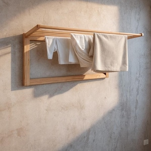 Könnte beinhalten: Ein wandmontierter Trockenständer aus Holz mit mehreren horizontalen Stangen und einem rechteckigen Rahmen. Weiße und beige Handtücher und Kleidung hängen auf dem Ständer. Der Ständer ist aus hellem Holz gefertigt und an einer strukturierten, hellfarbigen Wand befestigt.