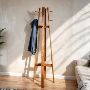 Puede incluir: Un perchero alto de madera con múltiples clavijas para colgar prendas. Una chaqueta azul cuelga del perchero. El perchero tiene un acabado de madera natural y un diseño minimalista, adecuado para entradas o dormitorios.