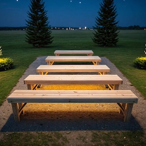 May include: An outdoor scene at night shows a series of wooden benches lit by warm lighting. The benches are arranged in a row along a gravel path, with green grass and trees in the background. The sky is dark with a few visible stars.