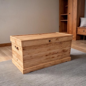 May include: A large wooden chest with a light brown finish. The chest has a rectangular shape with a flat top and metal handles on the sides. It is sitting on a gray rug.