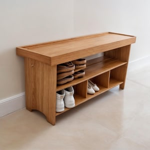 Puede incluir: Un zapatero de madera con un banco y estantes para guardar zapatos. El zapatero está hecho de madera marrón claro y tiene un acabado natural. Hay zapatos blancos y sandalias marrones en los estantes.