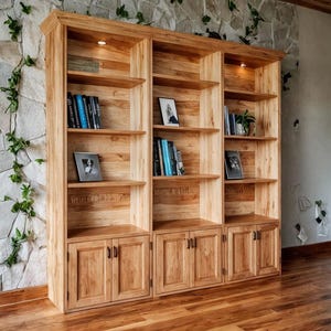 Op de afbeelding: Een grote, lichtgekleurde houten boekenkast met meerdere planken en kasten. De boekenkast is gevuld met boeken, ingelijste foto's en een kleine plant. De houtnerf is zichtbaar en het stuk heeft een warme, natuurlijke toon. De boekenkast staat tegen een stenen muur.