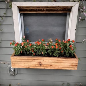 Puede incluir: Un jardinera de madera llena de vibrantes flores naranjas se encuentra debajo de una ventana. La jardinera está hecha de tablas de madera horizontales y está montada en una pared exterior gris. La ventana tiene un marco blanco y una pantalla.