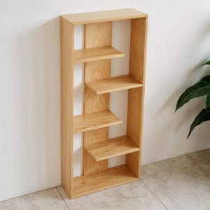 Puede incluir: Una estantería de madera clara con cuatro estantes. Los estantes están escalonados y soportados por paneles verticales de madera. La estantería está colocada contra una pared blanca, con una planta en el fondo.