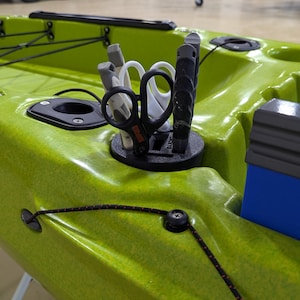 May include: A green kayak with a black and gray rod holder with fishing gear attached. The kayak has a blue storage box on the right side.