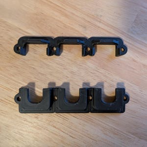 Può includere: Tre staffe in plastica nera stampate in 3D sono disposte su una superficie di legno. La fila superiore è composta da tre staffe individuali, ciascuna con una parte superiore curva e un piccolo foro. Sotto, tre staffe sono collegate.