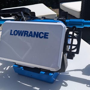 May include: A white and blue Lowrance fish finder with the brand name in blue lettering. The device is mounted on a blue and black frame. The image is taken outdoors on a sunny day.