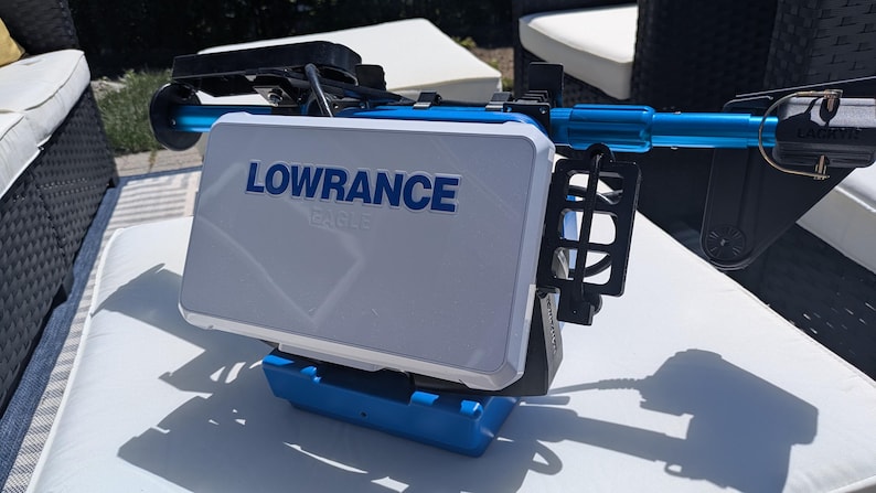 May include: A white and blue Lowrance fish finder with the brand name in blue lettering. The device is mounted on a blue and black frame. The image is taken outdoors on a sunny day.