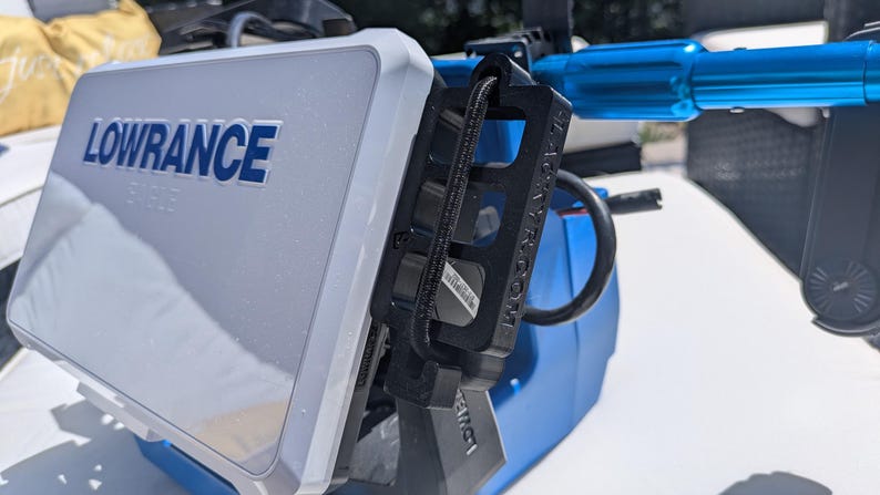 May include: A white and blue Lowrance fish finder with the brand name in blue lettering. The device is mounted on a blue and black bracket. The image is taken outdoors in daylight.