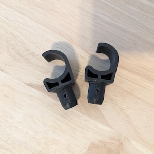 May include: Two black plastic hooks with a rounded top and a small hole in the center. The hooks are designed to be attached to a surface.