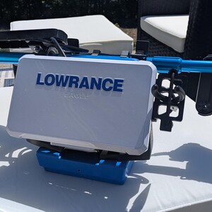 May include: A white and blue Lowrance fish finder with the brand name prominently displayed. The device is mounted on a blue and black frame. The image is taken outdoors on a white cushion.