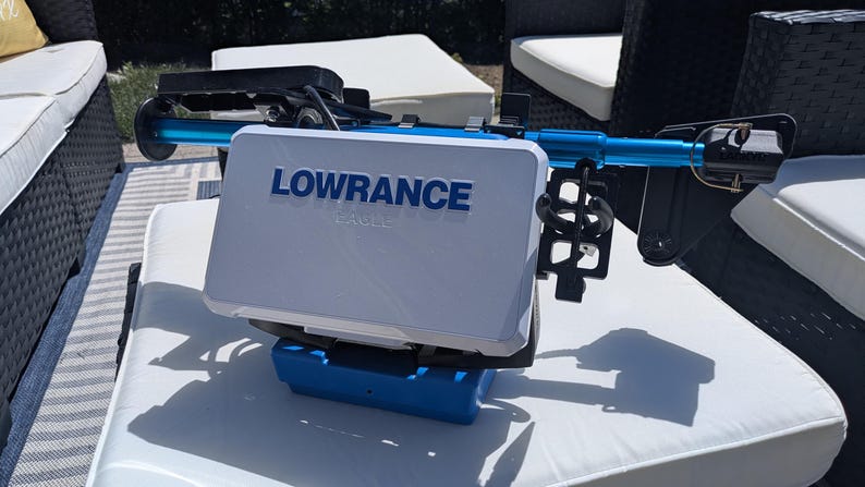 May include: A white and blue Lowrance fish finder with the brand name prominently displayed. The device is mounted on a blue and black frame. The image is taken outdoors on a white cushion.