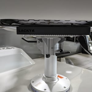 Puede incluir: Un pedestal de asiento de barco con una base rectangular negra con la etiqueta "LACKYR". El pedestal es plateado y tiene una base redonda. Una bandeja blanca con portavasos es visible en el fondo.