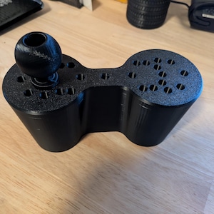 Puede incluir: Un organizador de escritorio negro impreso en 3D con una forma única. Cuenta con una pieza central de conexión con dos secciones redondeadas, cada una con múltiples agujeros pequeños. Un botón esférico negro está unido a un lado. El organizador está diseñado para guardar varios artículos.