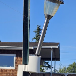 May include: A solar-powered outdoor lamp with a silver finish and a clear, ribbed plastic shade. The lamp is mounted on a green metal pole with a white plastic bracket. The background features a blue sky and a residential building.
