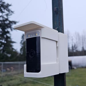 Puede incluir: Cámara de seguridad blanca con un protector de lluvia blanco montado en un poste de metal. La cámara tiene una lente negra y el texto "WYZE" es visible en la parte delantera.