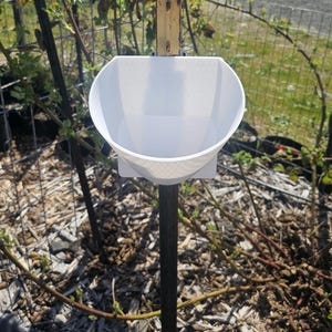 May include: A white plastic scoop attached to a black metal pole. The scoop is mounted to the pole with a wooden bracket. The pole is in a garden setting with brown mulch and green plants.