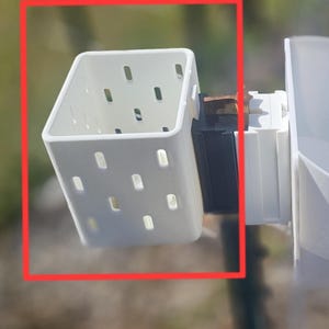 May include: A white plastic square container with rectangular holes is attached to a black and white structure. The container is angled, and the background is blurred green and brown.