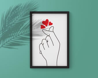 Op de afbeelding: Ingelijste kunstdruk met een handgebaar dat een hart vormt, met drie rode harten erboven. Het kunstwerk is een minimalistische zwart-wit lijntekening, geplaatst tegen een witte achtergrond in een zwart frame.