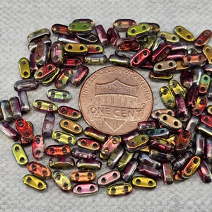 May include: A collection of small, iridescent glass beads in shades of red, gold, and green. The beads are shaped like small, elongated rectangles with two holes in each. The beads are scattered on a white surface, with a US penny in the center of the image.