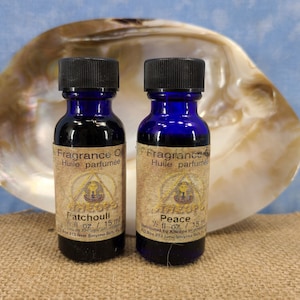 May include: Two small blue glass bottles with black caps, containing fragrance oil. The bottles are labeled "Patchouli" and "Peace" with the text "Fragrance Oil Huile parfumée" on each bottle. The bottles are sitting on a white and brown shell.