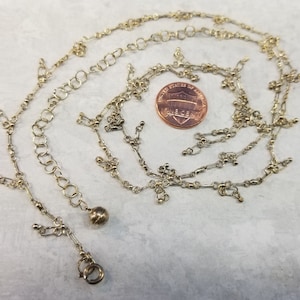 May include: A gold chain necklace with a small ball charm and a lobster clasp. The chain is made of multiple strands of small linked circles and has dangling charms.