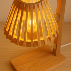 Puede incluir: Una lámpara de mesa de madera con una pantalla cónica hecha de listones de madera verticales. La lámpara tiene una base rectangular y un marco de soporte. La cálida luz de la bombilla brilla a través de la pantalla, creando un ambiente acogedor. La lámpara es de madera clara.