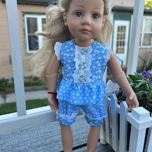 May include: A doll with long blonde hair and blue eyes wearing a blue polka dot outfit with white floral accents. The outfit includes a top with white lace trim and matching shorts. A white ribbon is in the doll's hair.