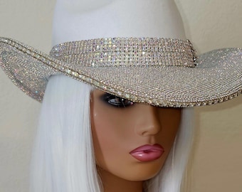May include: White cowboy hat with a wide brim and a sparkling rhinestone band around the crown and brim. The hat is displayed on a mannequin with white hair and a pale complexion. The hat is a glamorous accessory.