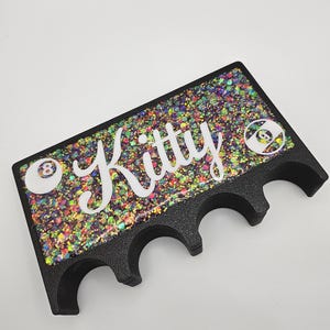 May include: Black pool cue rack with three slots. The rack has a glittery black surface with colorful glitter and the word "Kitty" written in white. The rack has two white 8-ball designs on either side of the word "Kitty".
