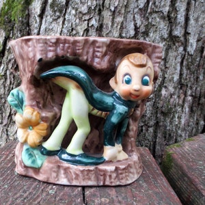 May include: A ceramic planter shaped like a hollow tree stump featuring a cartoon character with large blue eyes, a green tunic, and light-colored legs. A single yellow flower with green leaves is attached to the side of the brown textured stump.