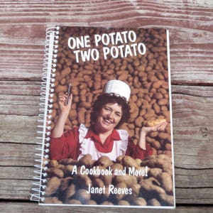 May include: A spiral-bound cookbook titled "ONE POTATO TWO POTATO" featuring a woman in a chef's hat and apron holding a potato and a knife. The cover includes the text "A Cookbook and More!" and the author's name, Janet Reeves.
