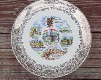 May include: A white ceramic plate with a gold rim and a decorative design. The plate features a map of New Hampshire with illustrations of various landmarks, including the Old Man of the Mountains, the State Capitol, and the Cogwheel Train on Mt. Washington. The text "NEW HAMPSHIRE THE GRANITE STATE" is printed in red.