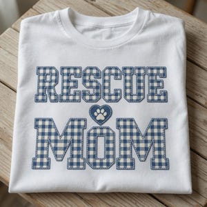 May include: A white short-sleeved t-shirt featuring the text "RESCUE MOM" in a blue and white gingham check pattern. Between the two words is a blue gingham heart containing a white paw print. The letters have a dark blue stitched outline.