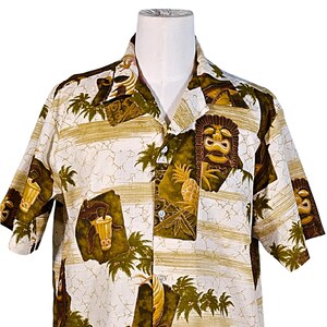 May include: A white short-sleeved button-down shirt with a tropical print featuring palm trees, tiki masks, and pineapple designs in shades of green, brown, and gold.
