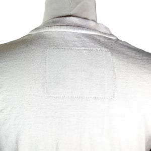 May include: A white turtleneck shirt, viewed from the back, with a rectangular patch detail on the upper back. The shirt is made of a soft, textured fabric, and the stitching is visible.