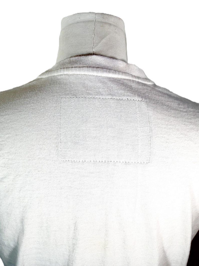 May include: A white turtleneck shirt, viewed from the back, with a rectangular patch detail on the upper back. The shirt is made of a soft, textured fabric, and the stitching is visible.