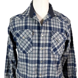 May include: A blue, gray, and white plaid flannel shirt with a dark blue collar. The long-sleeved shirt has two front pockets with silver snap closures and silver snaps down the front.