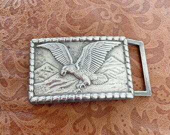 May include: A rectangular silver-colored belt buckle featuring a detailed relief of an eagle with outstretched wings soaring above a mountain range. The buckle has a beaded border and a silver-colored metal loop.