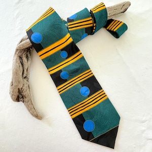 May include: A patterned necktie with a teal background, black stripes, yellow horizontal lines, and blue circles. The tie is knotted and rests on a piece of driftwood, showcasing a unique design.