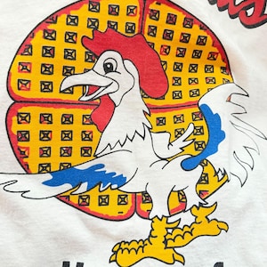 May include: White t-shirt with a colorful graphic. The design features a cartoon rooster with a red comb and blue accents, set against a yellow waffle background. The text "Waffthroats" arches above, with "House of chicken 'n Waff" below.