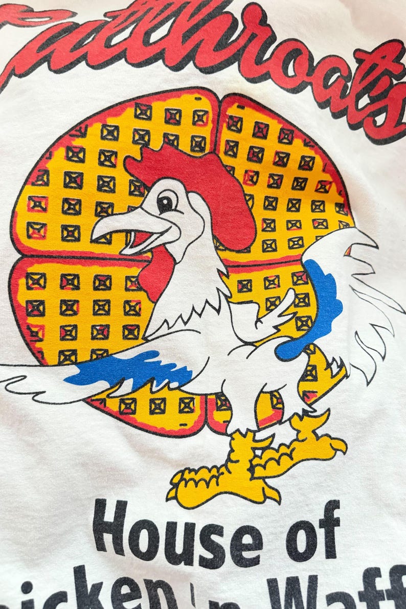 May include: White t-shirt with a colorful graphic. The design features a cartoon rooster with a red comb and blue accents, set against a yellow waffle background. The text "Waffthroats" arches above, with "House of chicken 'n Waff" below.